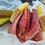 Large chunks of smoked meat inside two small pieces of bread. On the side you can see pickles.