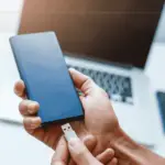 A hand holding a power bank, plugging in a usb cable in it. Behind, blurred, there is a laptop.