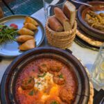 A small table with three dishes. A beef meatballs tagine, a plate with five cheese-filled filo triangles, and a lamb tagine.