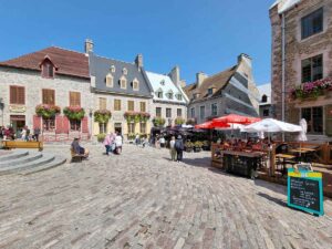 The main square in Quebec City, with typical stone buildings and cobbled stones on the ground. The windows of the houses have pink flowers in large pots underneath.