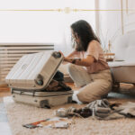 A woman packing a suitcase in her living room. There are electronic items on the floor next to her.