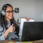 A woman wearing a grey shirt, black glasses and grey headphones, holding a paper in front of a black laptop, smiling at the camera.