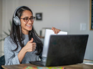 A woman wearing a grey shirt, black glasses and grey headphones, holding a paper in front of a black laptop, smiling at the camera.