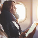 A close-up of a woman reading a book next to a woman sleeping, in the window seat.