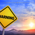A yellow sign with Warning written on black on it, against a mountain sunset.