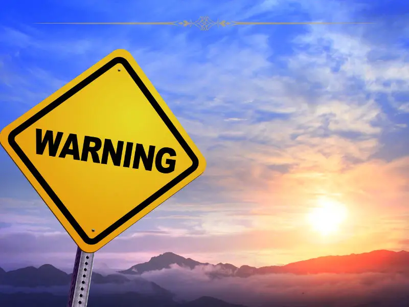 A yellow sign with Warning written on black on it, against a mountain sunset.