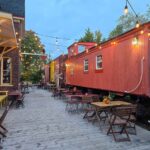 A red caboose in the train station, with tables and chairs in front and fairy lights going from it to the building.