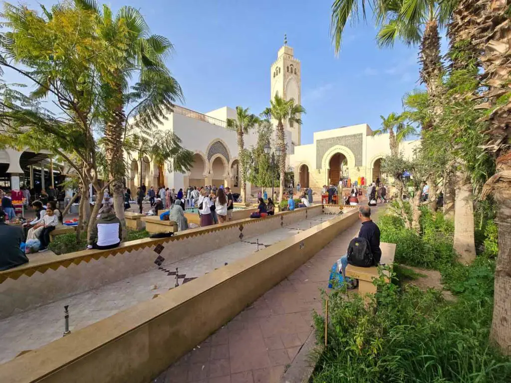 A mosque in the souk El Had in Agadir, with a water channel leading to it, surrounded by palm trees. There are a lot of people around. The sky is blue, without any clouds.