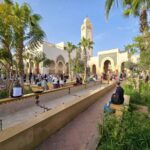 A mosque in the souk El Had in Agadir, with a water channel leading to it, surrounded by palm trees. There are a lot of people around. The sky is blue, without any clouds.