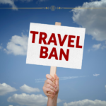 A hand holding a white sign with "travel ban" on it written in red letters, against the sky.