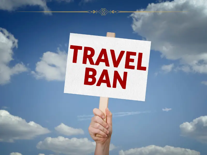 A hand holding a white sign with "travel ban" on it written in red letters, against the sky.