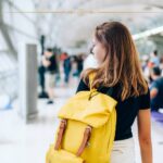A woman photographed from behind, at the airport, wearing a black tshirt, white trousers, and a yellow backpack.