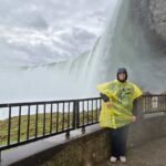 I am standing on the side of the falls, at the viewing platform from View behind the falls. I am wearing a yellow poncho over my jacket, which has the hoodie over my hear. I am smiling at the camera.