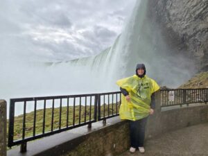I am standing on the side of the falls, at the viewing platform from View behind the falls. I am wearing a yellow poncho over my jacket, which has the hoodie over my hear. I am smiling at the camera.