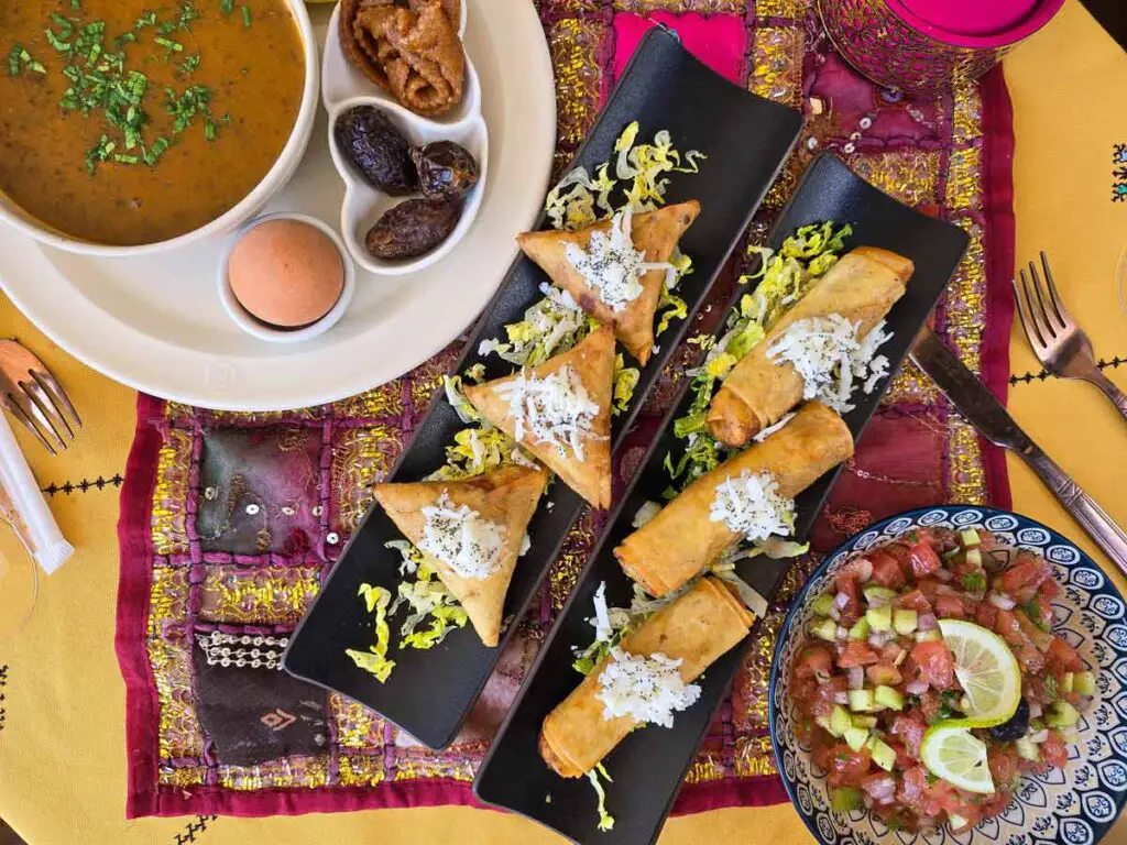 A bowl of soup in the top left part of the photo, two black long platters with pastries in the middle, and a bowl with a blue pattern filled with a fresh salad. The tablecloth is yellow and has a pink smaller one in the middle.
