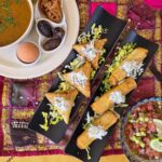 A bowl of soup in the top left part of the photo, two black long platters with pastries in the middle, and a bowl with a blue pattern filled with a fresh salad. The tablecloth is yellow and has a pink smaller one in the middle.