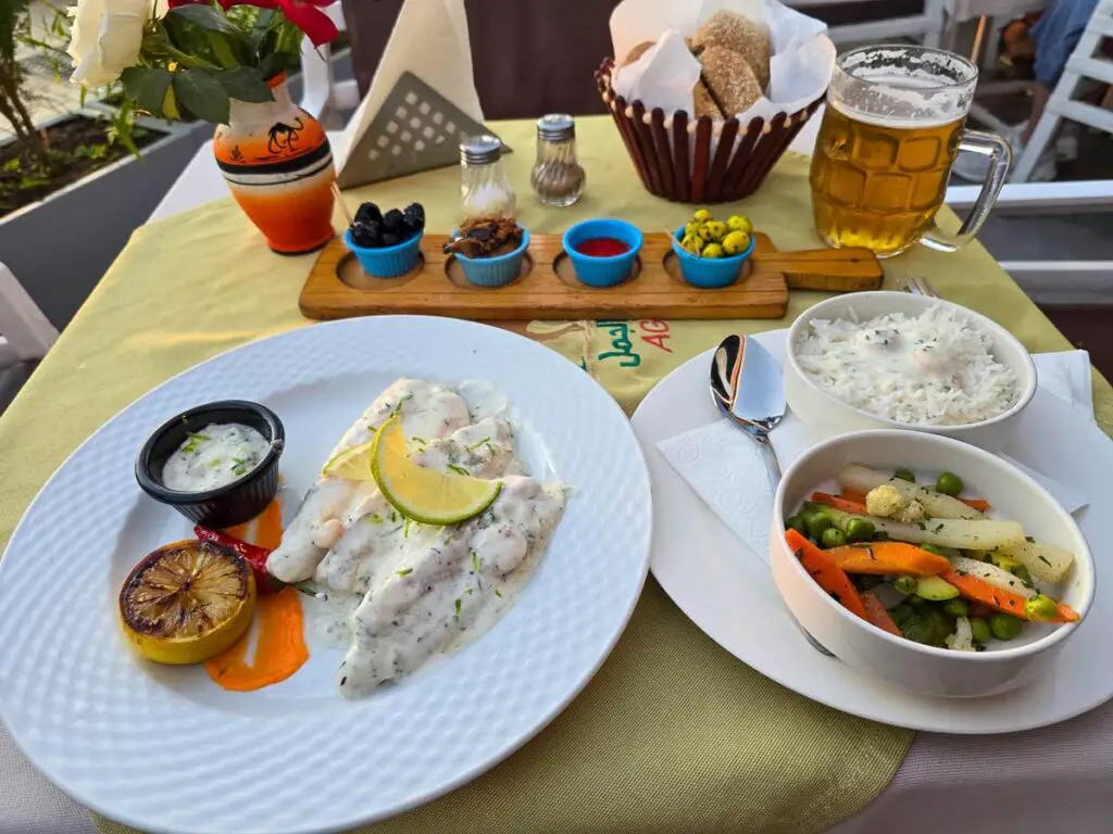 A white plate with a fish fillet on it covered in a white sauce, next to a black bowl with a white sauce in it and a grilled lemon. On the right there is another plate with a bowl filled with rice on it and another bowl filled with sauteed vegetables.