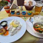 A white plate with a fish fillet on it covered in a white sauce, next to a black bowl with a white sauce in it and a grilled lemon. On the right there is another plate with a bowl filled with rice on it and another bowl filled with sauteed vegetables.
