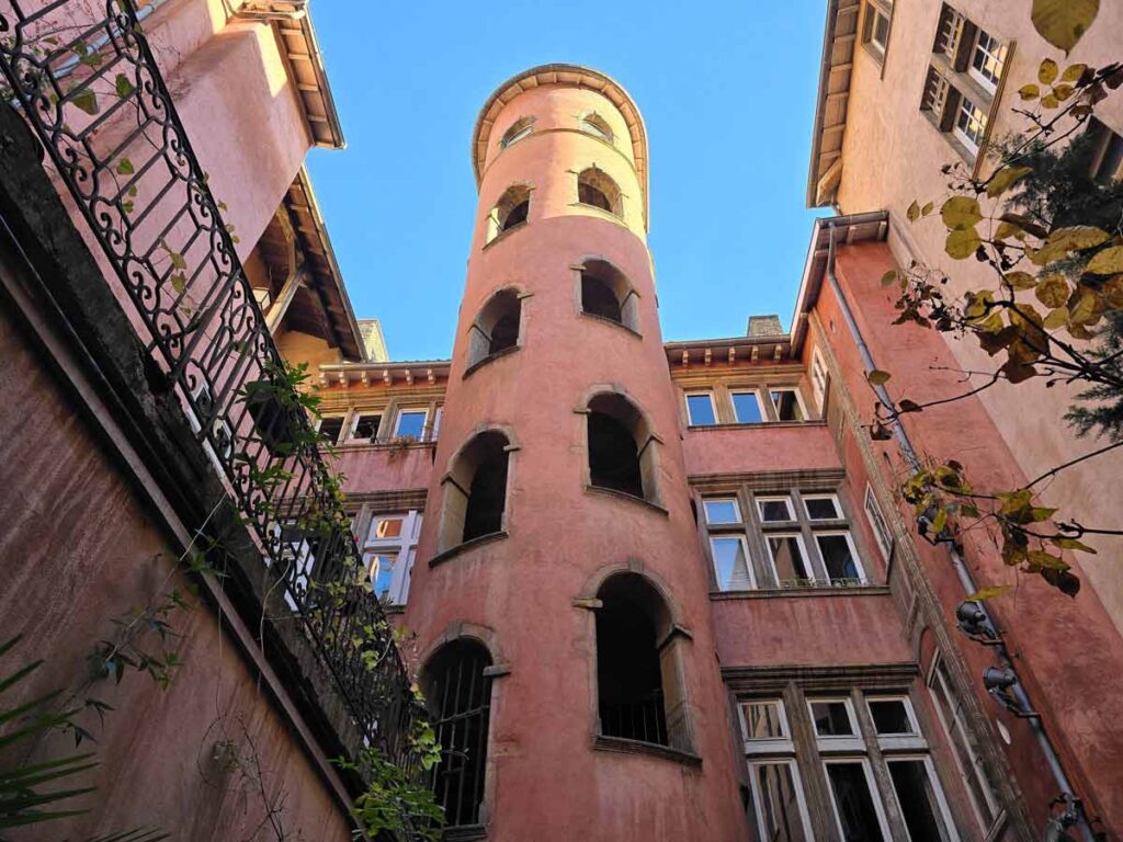 A courtyard in Lyon surrounded by pastel pink coloured buildings, including a round tower. The sky above is clear blue.