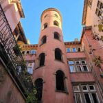 A courtyard in Lyon surrounded by pastel pink coloured buildings, including a round tower. The sky above is clear blue.