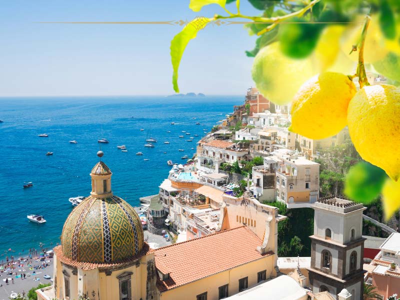 A view over Positano, with a lemon branch on the top left. You can see the sea and a bit of the beach underneath the cliffs on the left. The rest is the city descending down on the cliffs.