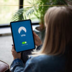 Travelling to Countries with Restricted Internet? Here’s How to Stay Connected and Informed The back of a woman sitting on a chair, holding a tablet with a blue screen and VPN written on it.