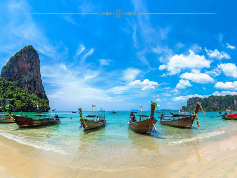 An exotic island in Thailand, with small boats on the golden sand and peaks covered in vegetation rising from the ocean.