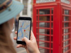 A close-up of a woman using Google Maps. She is standing next to a red phone box, typical for London.