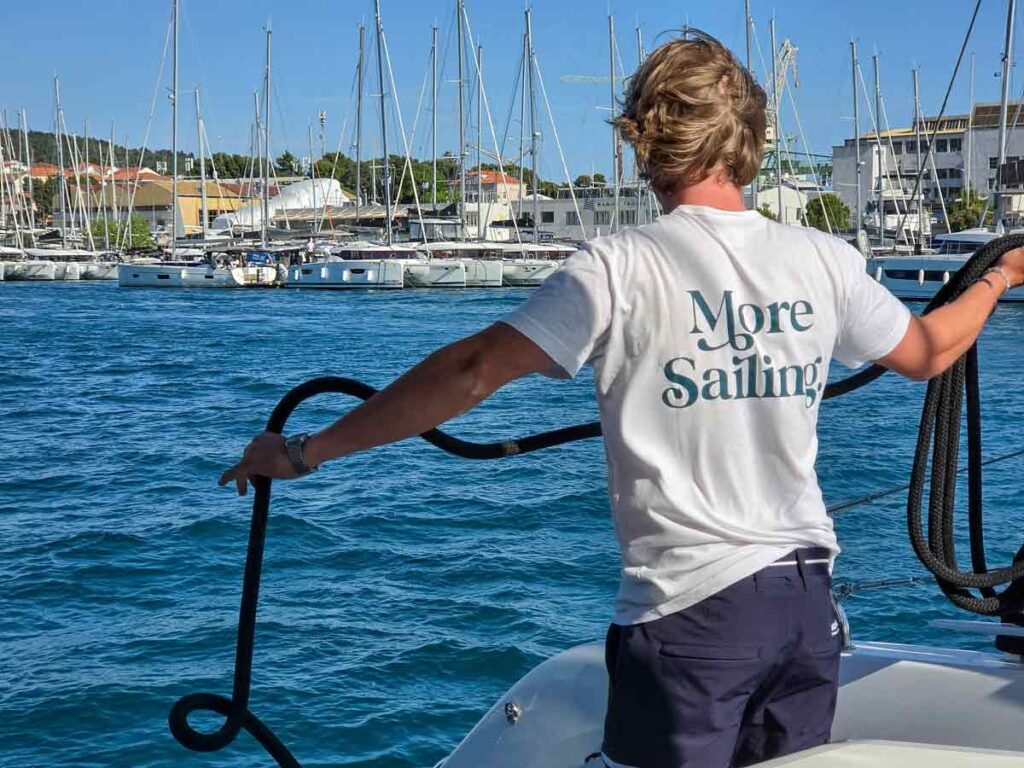 A man wearing a white tshirt with More Sailing written on it, holding rope, on a catamaran