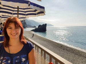 I am at a beach terrace in Italy, smiling at the camera. I am wearing a blue tshirt and I have red hair